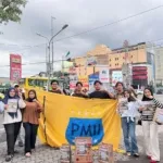 Seorang kader PMII memegang kotak donasi bertuliskan “Aksi Galang Dana Peduli Sumatera” sambil berdiri di pinggir jalan raya Gejayan, Yogyakarta, mengenakan jas almamater biru dan masker hitam.