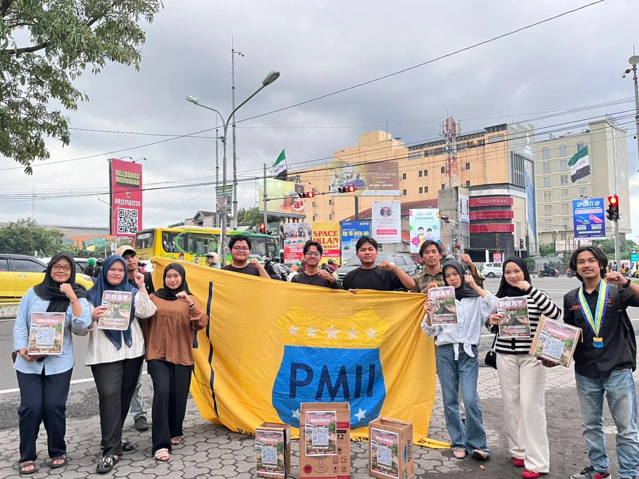 Seorang kader PMII memegang kotak donasi bertuliskan “Aksi Galang Dana Peduli Sumatera” sambil berdiri di pinggir jalan raya Gejayan, Yogyakarta, mengenakan jas almamater biru dan masker hitam.